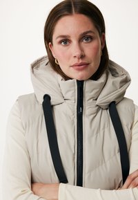 Gilet rembourré beige avec un col haut et une capuche, présentant des motifs de quilting diagonaux, une fermeture éclair noire et des sangles ajustables.