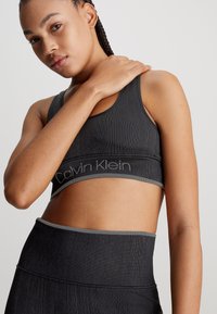 Czarny sportowy biustonosz w prążki z szarym wykończeniem, z widocznym logo Calvin Klein na dole. Modelka pozująca z jedną ręką na ramieniu.