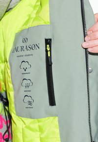 Giacca impermeabile verde con foderato giallo brillante, dotata di una tasca con zip e icone che indicano le caratteristiche impermeabile, traspirante e antivento.