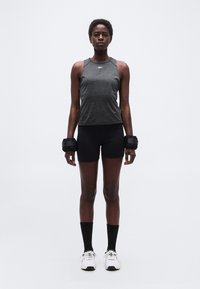 Γυναίκα που στέκεται, κοιτάζοντας μπροστά, φορόντας σκούρο αθλητικό τοπ χωρίς μανίκια, μαύρο σορτς, μαύρες κάλτσες, άσπρα αθλητικά παπούτσια και μαύρα βαράκια καρπού.