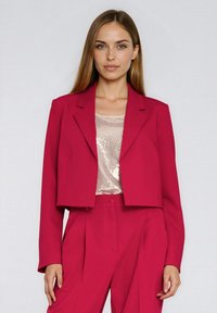 Fuksjowy krótki blezer o strukturalnym kroju, z wyciętymi klapami i długimi rękawami. Noszony na błyszczącym beżowym topie z cekinami, charakteryzujący się dopasowanym krojem.