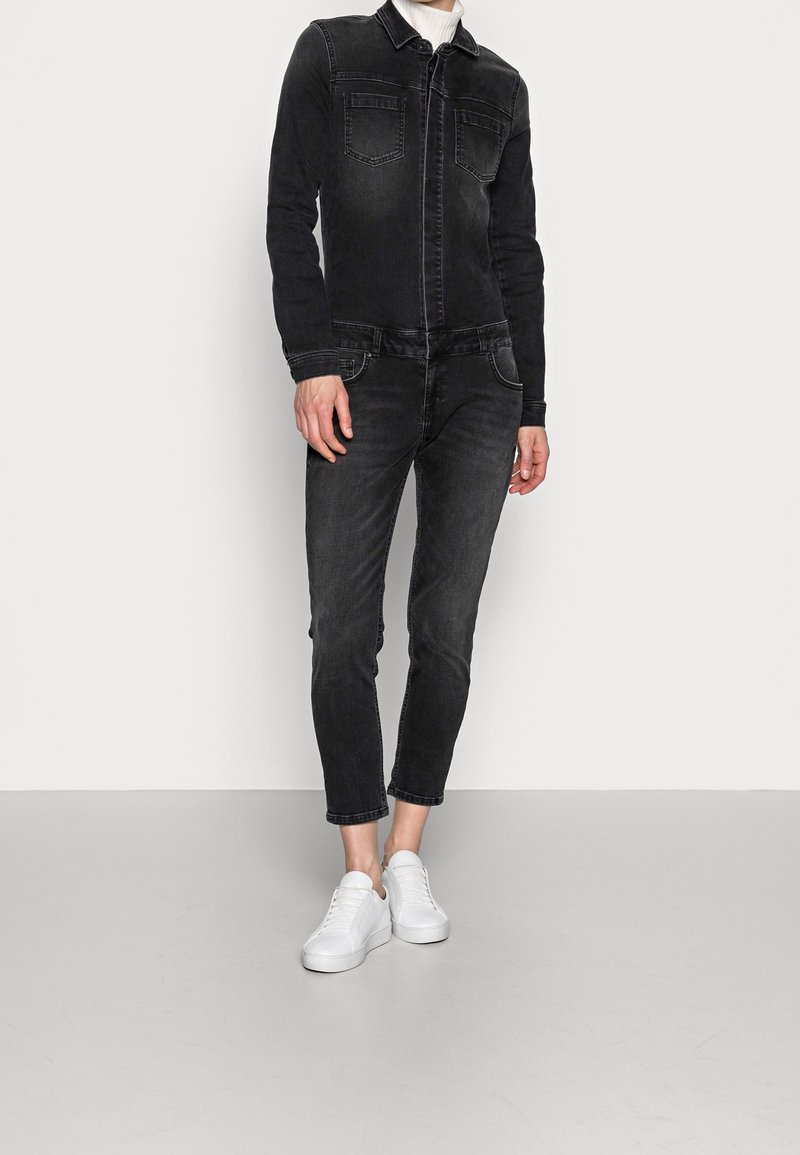 Personne portant une veste en jean noire et un pantalon avec un col roulé blanc et des baskets blanches, debout devant un fond uni.