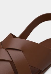 Καφέ δερμάτινο σανδάλι με φαρδιές, σταυρωτές λουρίδες και ένα μόνο λουράκι πίσω, σε απλό φωτεινό φόντο.