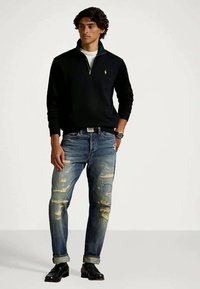 Ung man med svart tröja med kort dragkedja, slitna blå jeans, svarta skor, står med ena handen i fickan mot en enkel bakgrund.