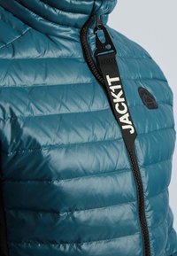 Teal puffjacka med horisontellt quiltat mönster, svarta dragkedjedetaljer och en logotypklistermärke. Har en hög krage och säker stängning.