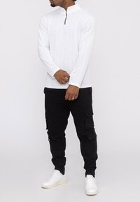 Haut à col zippé blanc à manches longues avec texture côtelée, associé à un pantalon cargo noir avec poches et des baskets blanches. Design minimaliste.