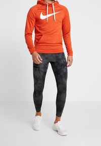 Sweat à capuche Nike orange avec logo blanc, leggings tie-dye gris et noir, et chaussures de sport blanches. La tenue présente un design décontracté et sportif.