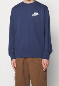 Person som bär en marinblå Nike-tröja med vit logotyp på bröstet och bruna cargo-byxor, stående mot en enfärgad ljus bakgrund.