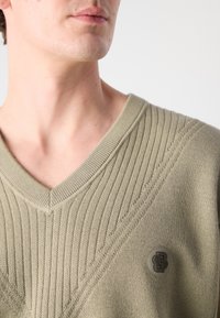 Sweater de malha em verde oliva claro com decote em V, apresentando um ribateado texturizado e um logotipo tonal no lado esquerdo. Tezzido suave, peso médio.