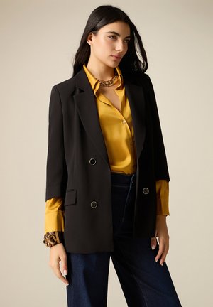 Frau trägt schwarzen doppelt geknöpften Blazer über senffarbenem Seidenhemd, dunkle Jeans, goldene Kette und klobiges goldenes Armband, steht vor beigem Hintergrund.