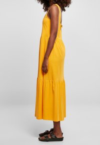 Żółta bawełniana sukienka maxi z warstwowym designem, regulowanymi ramiączkami i luźnym krojem. Połączona z ciemnymi sandałami.