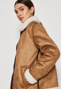 Cappotto in ecopelliccia color marrone, con un colletto in pelliccia bianca, chiusura a zip e tasche laterali. Presenta una vestibilità comoda e maniche lunghe.