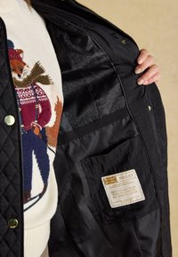 Main tenant une veste matelassée noire ouverte montrant une poche intérieure avec une étiquette Joules, portée sur un pull avec un renard sur le pull et un motif d'écharpe.