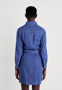 Denim skjortekjole i mellomblått med lange ermer, krage, belte i livet og knapper på mansjettene. Har vertikale sømmer og lommer.