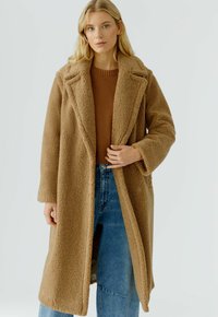 Blonde vrouw in een lange beige teddyjas over een bruine trui en blauwe jeans voor een eenvoudige lichte achtergrond.