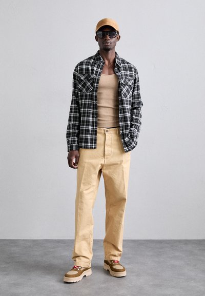 Ternet sort og hvid flannel skjorte, beige tank top, beige løse bukser, brune og beige sneakers, gul kasket, mørke solbriller.