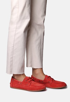 Personne portant un pantalon court écru et des mocassins rouges en daim, debout sur un fond clair uni.