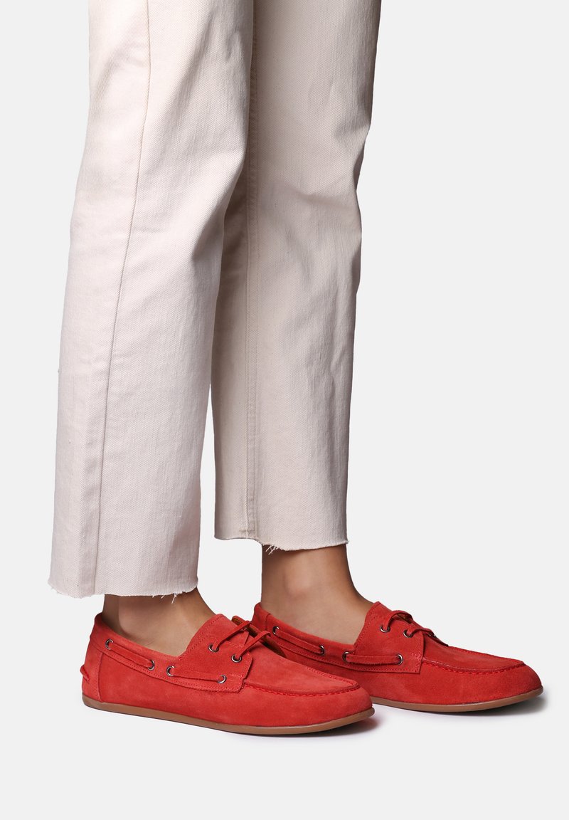 Person iført off-white cropped bukser og røde mokkasinstillet sko i ruskind, stående på en ensfarvet lys baggrund.