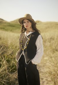Un gilet en laine noir avec des bords blancs, superpos�é à une chemise blanche brodée. Le modèle porte un chapeau beige et un gros collier jaune. Fond en herbe verte.