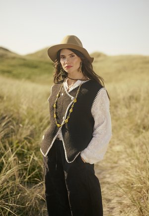 Un gilet en laine noir avec des bords blancs, superposé à une chemise blanche brodée. Le modèle porte un chapeau beige et un gros collier jaune. Fond en herbe verte.