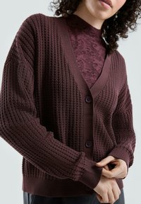 Burgundowy sweter kardigan z dzianiny o fakturowanym wzorze, dekoltem w serek, ściągaczami na rękawach i dwoma guzikami, noszony na dopasowanym topie z półgolfem.