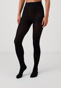 Person iført næsten helt sorte strømpebukser, stående med fødderne lidt fra hinanden på en lys grå gulv og baggrund.