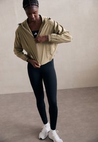 Kvinne i sorte leggings, hvite joggesko og beige jakke som drar opp glidelåsen i et minimalistisk innendørsmil med beige vegger og grått gulv.