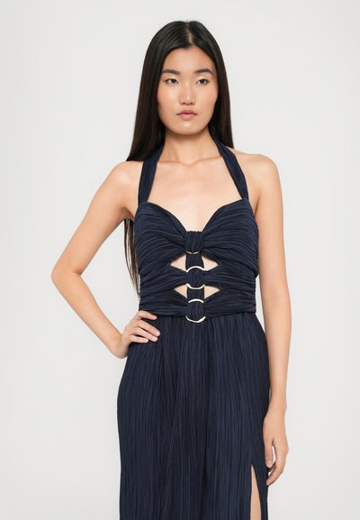 Navy blue pleated dress with halter neck. Features a knotted front and cutouts adorned with gold rings. Flared skirt with side slit.