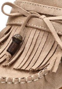 Mocasines de ante beige con detalles de flecos, un lazo de cuero atado en forma de moño, y un adorno de cuentas de madera cerca de la parte delantera.