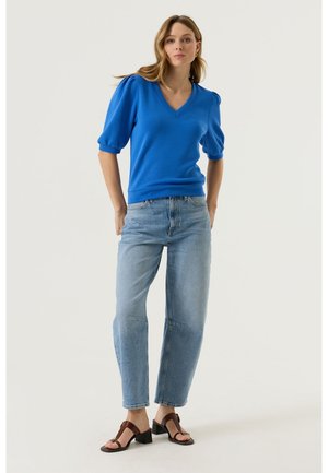 Donna in piedi con indosso una maglia blu con scollo a V e maniche a sbuffo, jeans azzurri chiari e sandali con tacco marroni, su uno sfondo semplice.