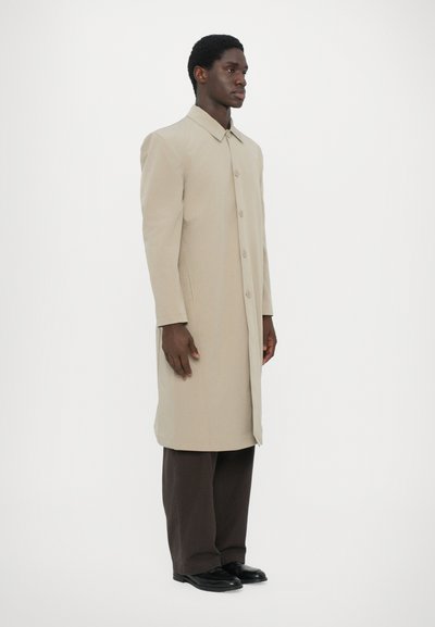 Beige long coat made from a smooth fabric, featuring a pointed collar and front button closure. Model wears dark brown wide-leg pants and shoes.