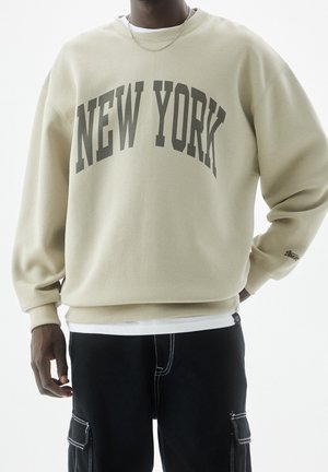 Personne portant un sweat-shirt beige avec "NEW YORK" en grandes lettres sombres, associé à un pantalon cargo noir avec des coutures blanches.