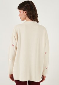 Maglione lavorato a maglia di colore crema con una vestibilità morbida, caratterizzato da ricami a forma di fiocco rosso sulle maniche e un design del colletto semplice.