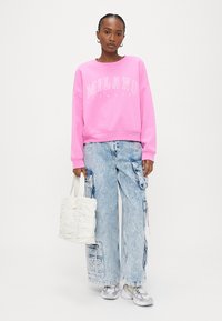Frau trägt rosa Sweatshirt mit Aufschrift "Milano Italia", kariotte Cargo-Jeans mit Acid-Waschung, silberne Turnschuhe und hält eine weiße Ruched-Tasche.