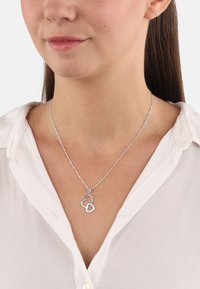 Silverhalsband med ett dubbelhärtpandent; en hjärta är konturerad och den andra är fylld med rhinestones. Modellen bär en vit skjorta.