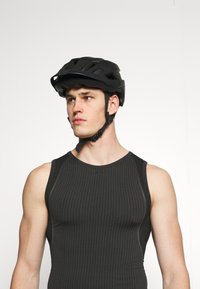 Czarny kask rowerowy z otworami wentylacyjnymi, regulowanymi paskami i eleganckim designem. Model nosi dopasowany czarny top na ramiączkach o teksturowanej tkaninie.
