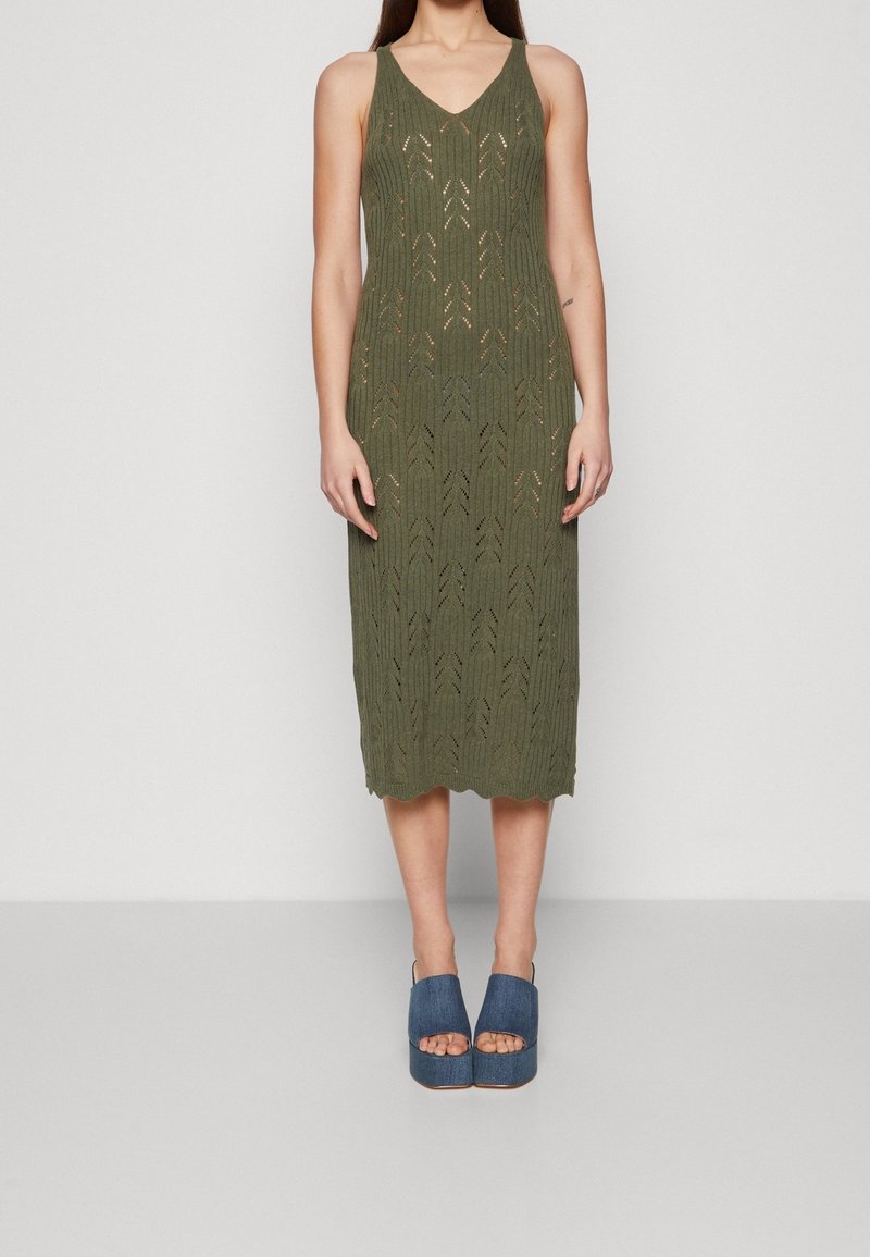 Femme portant une robe midi en maille sans manches vert olive avec un col en V et des trous décoratifs, associée à des sandales à plateforme bleues à bout ouvert.