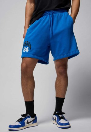 Uomo che indossa pantaloncini Jordan di un brillante blu con il numero "84" stampato sulla gamba sinistra, calzini neri a costine e scarpe Nike bianche, blu e nere.