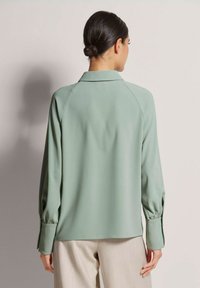 Helle grüne Bluse mit glatter Textur, langen Ärmeln und einem Kragen. Verfügt über geraffte Manschetten und ein lässiges Design.