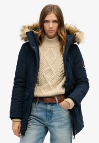 Parka azul marino con capucha forrada de piel, con cierre de cremallera y botones. Llevada sobre un jersey de punto color crema y jeans azules, acentuada por un cinturón marrón.