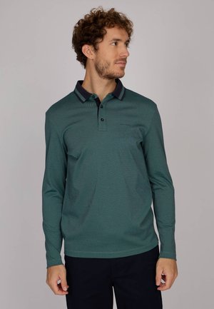 Homme avec des cheveux bouclés portant un polo à manches longues vert avec un col sombre, regardant vers sa droite sur un fond uni.