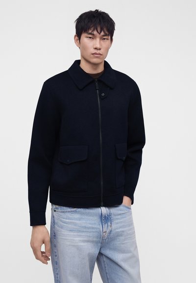 Jeune homme portant une veste noire zippée avec des poches à rabat et un jean bleu clair, debout devant un fond clair uni.