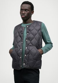 Πλισέ γκρι γιλέκο με στρογγυλή λαιμόκοψη και κουμπιά με κούμπωμα, διαθέτοντας μεγάλες τσέπες και λεία υφή, τοποθετημένο πάνω από μπλούζα με μακριά μανίκια σε απόχρωση τυρκουάζ.