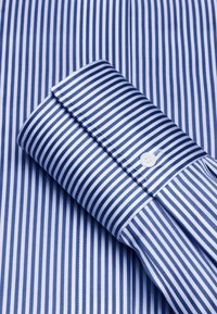 Camicia a righe blu e bianche con maniche arrotolate, dotata di chiusura a bottoni e texture liscia e croccante. Dettagli di cucitura chiari visibili.