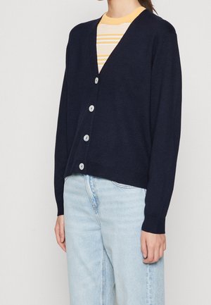 Femme portant un cardigan bleu marine boutonné par-dessus une chemise rayée jaune et blanche avec un jean bleu clair, debout devant un fond uni.