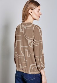 Brun tröja med abstrakta beige mönster, trekvartslånga ärmar, rundad halsringning och mjuk textur, matchad med mörk denimjeans.
