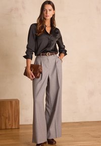 Camicia di raso nera con colletto, pantaloni grigi plissettati a vita alta, cintura in pelle marrone e borsa marrone testurizzata. Sfondo in legno.