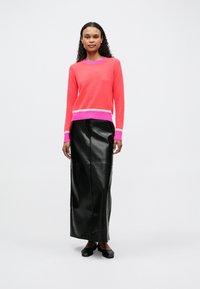 Mujer con cabello rizado que lleva un suéter rosa y coral, una falda maxi de cuero negra y zapatos negros, de pie contra un fondo blanco.