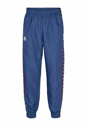 Mand iført blå Kappa træningsbukser med hvide logostriber, navy sneakers med hvide snørebånd og en rød og hvid jakke.