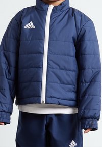 Námorně modrá péřová bunda, prošívaný design, bílý zip, vysoký límec, pružné manžety, s kontrastním bílým logem Adidas na hrudi.
